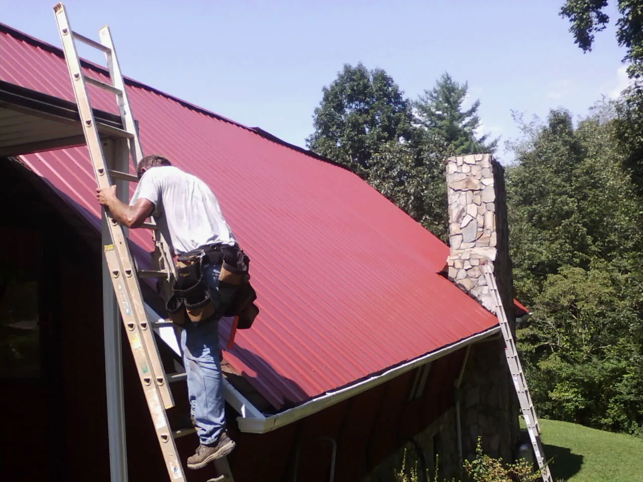 Roofer on a ladder beside a red metal roof and stone chimney — Soffit & Fascia Repair by Roofing Brothers in Tolland